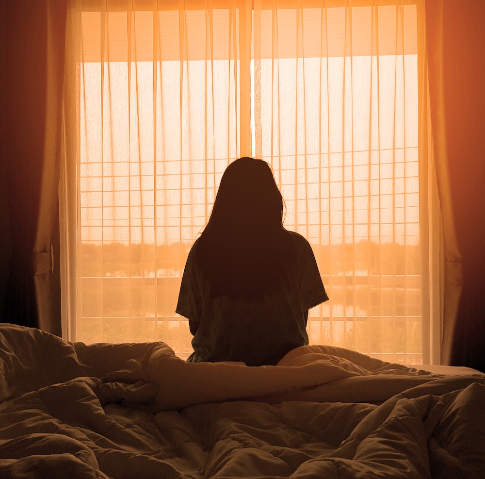 silhouette of woman sitting on the bed beside the windows with sunlight in the morning